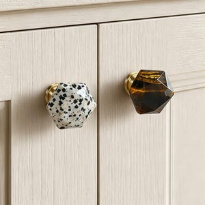 May include: Close-up of two cabinet knobs. One is white with black speckles, the other is dark amber. Both have a faceted, geometric design and gold-toned bases. They are attached to a light-coloured cabinet. Ideal for home decor.
