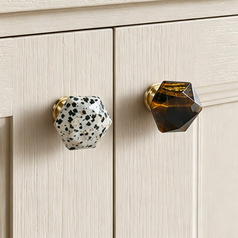 May include: Close-up of two cabinet knobs. One is white with black speckles, the other is dark amber. Both have a faceted, geometric design and gold-toned bases. They are attached to a light-coloured cabinet. Ideal for home decor.