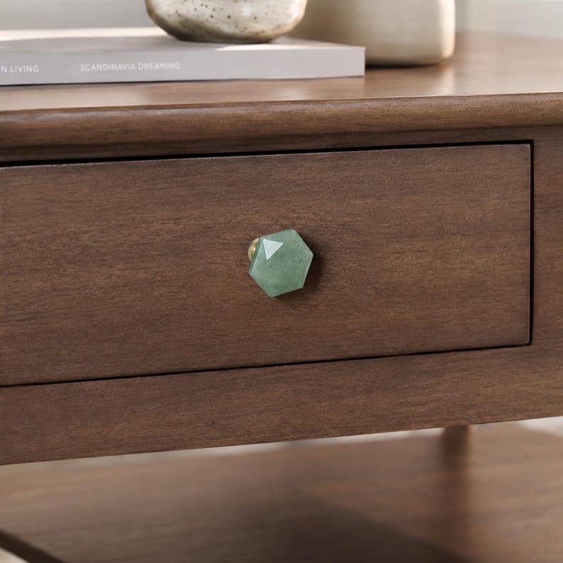 May include: A close-up shot of a wooden drawer featuring a green, faceted gemstone knob. The drawer is part of a larger piece of furniture, possibly a bedside table or chest of drawers. The knob is hexagonal with a gold base.