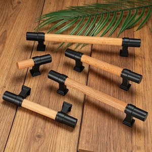 Puede incluir: Una colección de herrajes para muebles con tiradores cilíndricos de madera clara, rematados con tapas metálicas en negro mate y bases de montaje cuadradas. El conjunto incluye un pomo pequeño y tres tiradores de cajón de distintas longitudes.