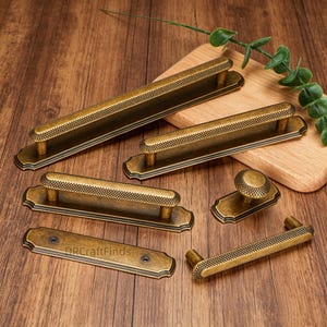 Puede incluir: Un surtido de herrajes de gabinete de latón antiguo, que incluyen manijas y un pomo. Las manijas tienen un agarre texturizado con patrón de diamantes y están montadas en placas traseras rectangulares. Los herrajes se exhiben sobre una superficie de madera.