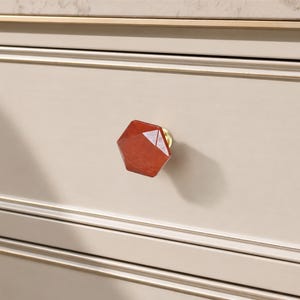 May include: A close-up of a red, geometric wooden drawer knob with a brass base. The knob is attached to a light beige drawer with gold trim. The design is modern and adds elegance to the furniture.