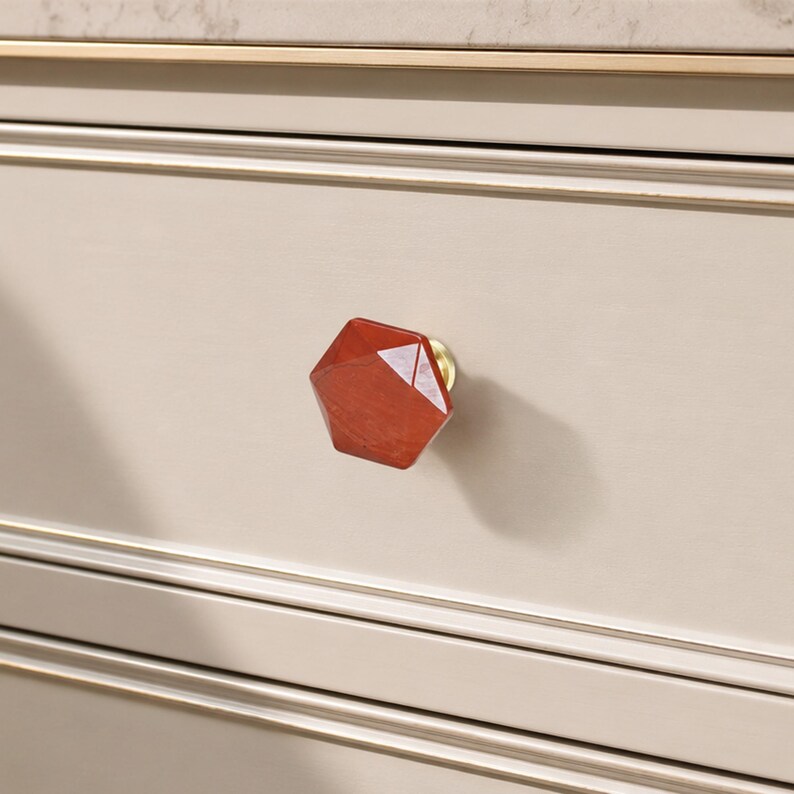 May include: A close-up of a red, geometric wooden drawer knob with a brass base. The knob is attached to a light beige drawer with gold trim. The design is modern and adds elegance to the furniture.