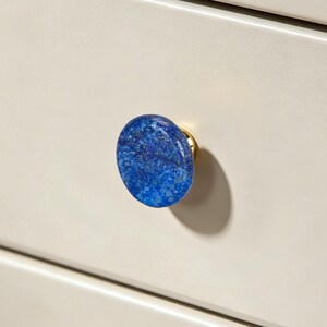 Może przedstawiać: Okrągła gałka meblowa z polerowaną powierzchnią z kamienia lapis lazuli w kolorze głębokiego błękitu z naturalnymi inkluzjami mineralnymi oraz złotymi okuciami, zamontowana na gładkim, jasnobeżowym froncie szuflady z poziomym łączeniem.