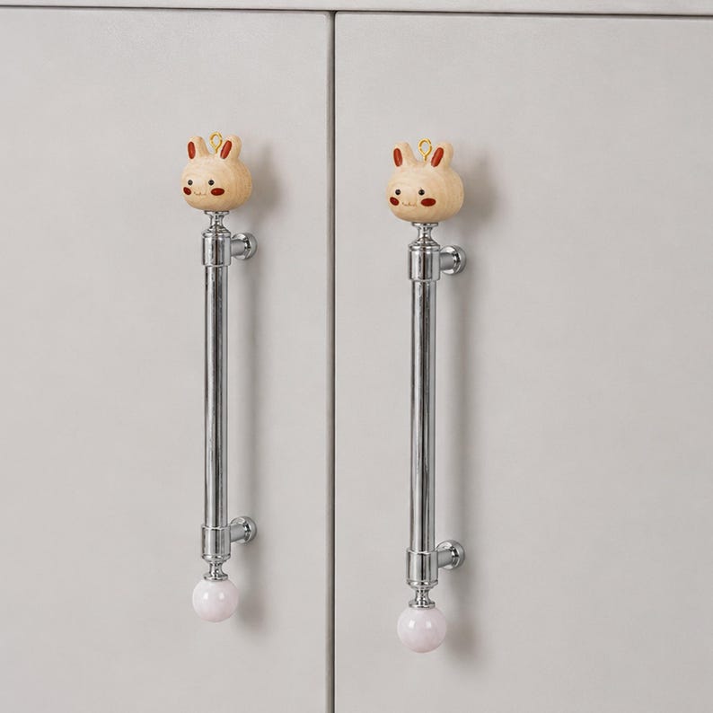 May include: Two vertical cabinet handles featuring a polished chrome finish. Each handle is topped with a light-colored wooden rabbit head ornament with painted facial features and a small gold loop, ending with a smooth white spherical base.