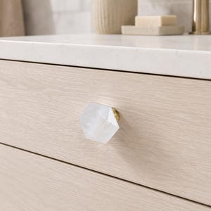 May include: A close-up of a light wood drawer with a clear, faceted, hexagonal knob. The knob has a gold-coloured base. The drawer is part of a light-coloured wooden cabinet. The background includes a white countertop and bathroom accessories.