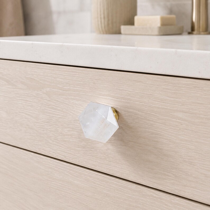 May include: A close-up of a light wood drawer with a clear, faceted, hexagonal knob. The knob has a gold-coloured base. The drawer is part of a light-coloured wooden cabinet. The background includes a white countertop and bathroom accessories.