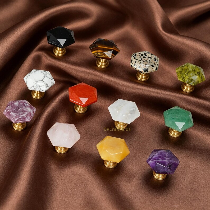 May include: A selection of gemstone cabinet knobs in a range of colours, including black, brown, white, red, clear, green, pink, yellow, and purple. Each knob has a faceted, hexagonal shape and a brass base. The knobs are displayed on a brown satin surface.