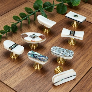 Op de afbeelding: Een verzameling decoratieve kastknoppen met verschillende ontwerpen. De knoppen hebben witte, iriserende en patroonoppervlakken, geplaatst op goudkleurige bases. De vormen omvatten rechthoekige, ovale en gebogen ontwerpen, met een reeks texturen en kleuren.