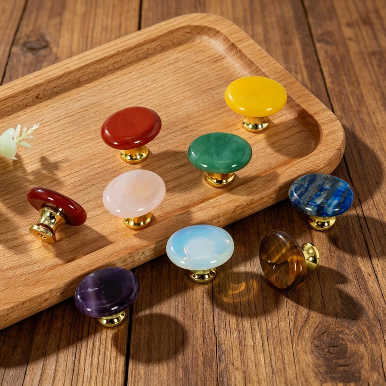 May include: A collection of nine round cabinet knobs featuring smooth, polished stone tops in various colors including yellow, red, green, blue, white, and purple, each attached to a shiny gold-toned metal base for furniture attachment.