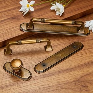Può includere: Una collezione di ferramenta in ottone anticato che include una lunga maniglia per cassetto con piastra, una maniglia più piccola, un pomello rotondo per armadietto su piastra rettangolare e una piastra rettangolare lunga separata con due fori.