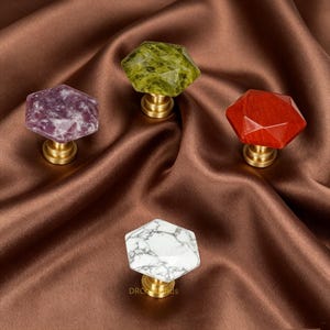 May include: Four decorative cabinet knobs with faceted, gemstone-like tops in purple, green, red, and white marble. Each knob is mounted on a brass base. The knobs are arranged on a brown satin fabric.