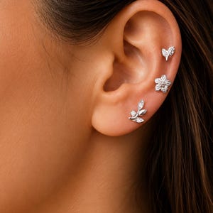 Könnte beinhalten: Nahaufnahme eines Ohrs, das mit drei silbernen Ohrsteckern geschmückt ist. Die Ohrringe haben ein Blatt-, Blumen- und Herzdesign, jedes mit kleinen, funkelnden Steinen verziert. Das Ohr ist vor einem warmen Hautton platziert.