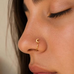 Può includere: Primo piano di un naso adornato con due gioielli. Un piccolo piercing al naso a forma di mezzaluna dorata è posto sopra un anello al naso dorato. Il testo "Sterling Silver 925" e "Hypoallergenic" sono visibili.