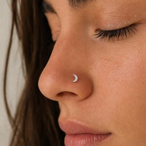 Può includere: Primo piano di un naso con un piccolo piercing d'argento a forma di mezzaluna. La luna è adornata da piccole pietre scintillanti. La pelle è liscia e lo sfondo è sfocato.