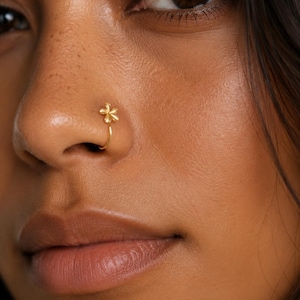 Può includere: Un anello da naso color oro caratterizzato da un piccolo design a fiore con cinque petali. Il gioiello è formato da una sottile fascia curva che poggia sulla narice, mostrando una finitura metallica liscia e un delicato elemento decorativo floreale.