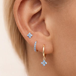 Könnte beinhalten: Nahaufnahme eines Ohrs, das mit drei goldenen Ohrringen geschmückt ist. Einer ist ein Stecker mit einem vierblättrigen Blumenmuster, ein anderer ist ein kleiner Reifen mit blauen Steinen, und der dritte ist ein hängender Reifen mit einem Blumenanhänger.