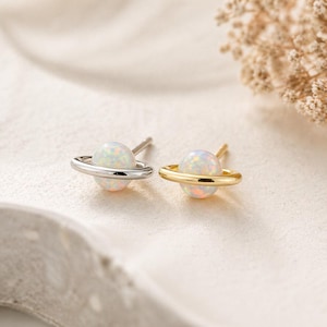 Può includere: Un paio di orecchini a lobo con un design a forma di pianeta. Uno è argentato, l'altro dorato. Ognuno ha una sfera bianca, simile all'opale, circondata da una fascia metallica. Gli orecchini sono su uno sfondo neutro.