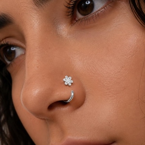 Puede incluir: Un aro de nariz plateado con un pequeño dije de flor de seis pétalos, con piedras claras y brillantes incrustadas en cada pétalo. La joya consiste en un aro circular que pasa a través de la fosa nasal y se conecta con la flor decorativa.