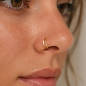 Op de afbeelding: Close-up van een gezicht met een gouden dubbele neusring. De ring bestaat uit twee dunne gouden bandjes. De huidskleur is licht en de achtergrond is wazig.