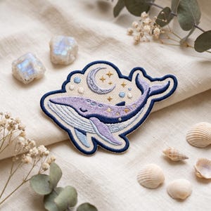 May include: An embroidered patch featuring a light purple whale with a cream-colored belly, surrounded by a dark blue border. Above the whale, a crescent moon and small gold stars are stitched against a cream background with light blue circles.