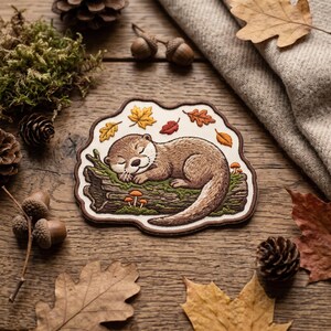 May include: An embroidered patch featuring a brown otter sleeping on a mossy log with small orange mushrooms. The patch is surrounded by scattered autumn leaves in shades of yellow, orange, and red, all set against a cream-colored background.