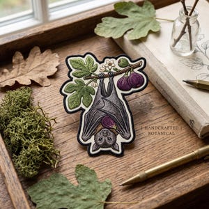 May include: An embroidered patch featuring a grey bat hanging upside down from a fig tree branch with green leaves, small white flowers, and two purple figs. The patch has a black border and detailed stitching throughout the design.