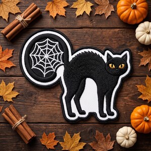 May include: An embroidered patch featuring a black cat with arched back and yellow eyes, with a white spiderweb design on its curled tail. The patch has a thick black and white border and is displayed against a dark wooden background.