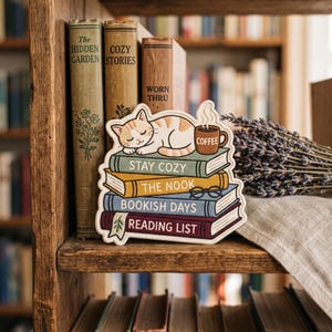 May include: A decorative sticker featuring a sleeping orange tabby cat on a stack of four books. The books are labeled STAY COZY, THE NOOK, BOOKISH DAYS, and READING LIST. A brown mug labeled COFFEE and black glasses are also illustrated on the books.