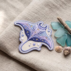 May include: An embroidered patch shaped like a manta ray, featuring shades of purple, blue, and cream. The design includes small gold star accents and circular patterns. The patch has a thick, raised border and is displayed on a textured fabric surface.