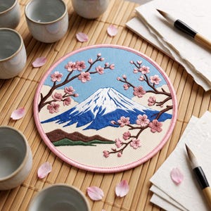 May include: A circular embroidered patch featuring a detailed landscape of Mount Fuji with a snow-capped peak, dark blue slopes, and green foothills. The scene is framed by brown branches with pink cherry blossoms against a light blue sky.