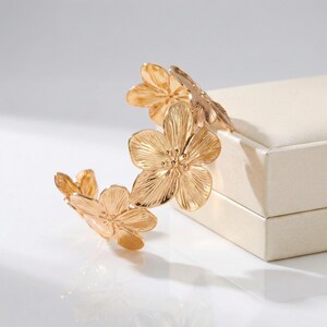 May include: A gold-coloured floral cuff bracelet. The bracelet features multiple detailed flower designs. The flowers are arranged in a curved shape, with the petals and centres clearly defined. The bracelet is displayed on a white surface.