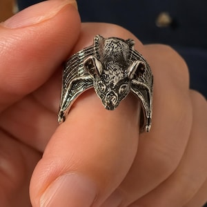 Puede incluir: Un anillo de metal en tono plateado que presenta un diseño detallado y esculpido de un murciélago. El murciélago se muestra con las alas extendidas formando la banda del anillo, con detalles de pelaje texturizado en la cabeza y el cuerpo.