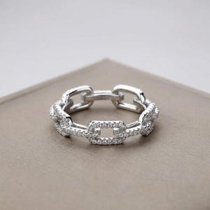 Könnte beinhalten: Ein silberner Ring mit einem Kettenglied-Design. Der Ring hat eine Reihe von rechteckigen Gliedern, von denen einige mit kleinen, klaren Edelsteinen besetzt sind. Der Ring ist vor einem neutralen Hintergrund platziert.