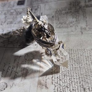 May include: A silver pendulum with a clear quartz crystal point. The pendulum has a detailed wing design and small rose accents. The background is a vintage paper with script.