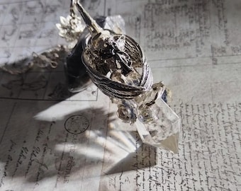 May include: A silver-toned pendulum with a clear quartz crystal point. The pendulum features a detailed wing design and small rose accents. The background is a vintage paper with script.