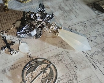 May include: A silver-toned pendant with an owl design, a faceted crystal, and a circular charm with a crescent moon and star motif. The pendant is attached to a chain with a cross and white beads. The background is a vintage paper.