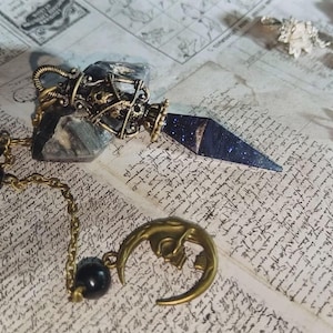 Pode incluir: Imagem detalhada de um pingente e colar com tema celestial. O pingente apresenta uma pedra azul escura salpicada de estrelas com uma caixa de metal cor de bronze. Um pingente de lua crescente e uma conta preta também são visíveis, conectados por uma corrente.