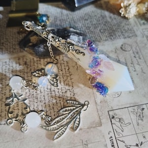 May include: A decorative crystal pendant with a silver-toned metal top and chain. The crystal is adorned with blue and purple floral accents. Additional charms include a leaf and a winged orb.