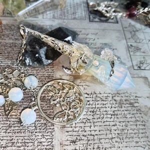 Puede incluir: Un colgante plateado con una punta de cristal y detalles decorativos. El colgante está unido a una cadena con cuentas blancas en forma de orbe y un dije circular con un diseño floral. El cristal es de color azul pálido.