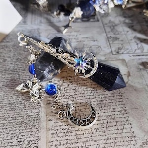 Pode incluir: Uma presilha de cabelo com tema celestial com uma lua crescente prateada, raios de sol e detalhes em pedras preciosas azuis. A presilha é adornada com pingentes pendurados, incluindo uma esfera azul e uma borboleta prateada.