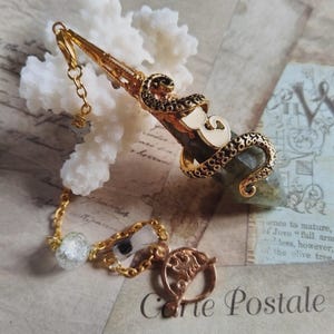 May include: A gold-toned metal pendulum featuring a dark, faceted stone point wrapped in an octopus tentacle design. The pendulum is attached to a gold chain with a clear bead, a small rectangular charm, and a circular charm with an embossed design.