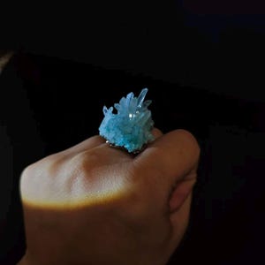 May include: A unique ring featuring a cluster of light blue crystals. The crystals are clustered together, creating a textured, eye-catching design. The ring's band appears to be silver-toned, complementing the cool tones of the crystals. The ring is set against a dark background.