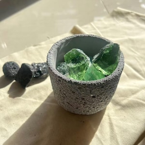 May include: A small, grey, textured concrete bowl filled with bright green glass pieces. Several dark grey, porous stones are scattered nearby. The bowl sits on a beige fabric surface, with sunlight casting a shadow.