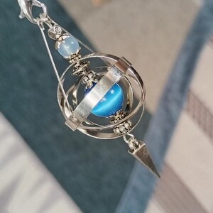 May include: A silver pendant with a blue orb within a spherical cage. The pendant has a light blue bead at the top and a pointed base. The design is detailed, with a focus on light and shadow. The pendant's design is intricate and detailed.