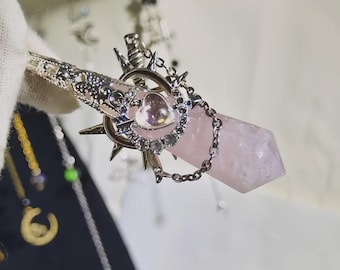 May include: A silver-toned wand with a pink crystal point. The wand features ornate metalwork, including a heart-shaped embellishment and a chain. The crystal is a pale pink color.