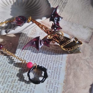May include: A gold-toned metal pendant featuring a dark purple bat wing design and a central crystal point. The piece is attached to a gold chain adorned with a small pink bead and a black circular ring charm, resting on a printed paper surface.