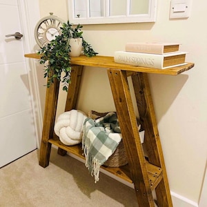 May include: A wooden console table with an A-frame design. The table has a lower shelf with a woven basket, a white knot pillow, and a green and white checkered throw. The top shelf displays a clock, a potted plant, and two books.