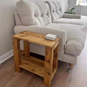 May include: A small, wooden side table made from reclaimed wood, with a lower shelf. The table displays natural wood grain and knots. A set of square coasters sits on top. The table is positioned next to a light-coloured, upholstered sofa.