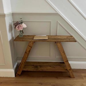 May include: A reclaimed wood console table with a natural finish. The table features a rectangular top and angled legs, with a lower shelf. A small vase of flowers and a brown envelope are on the tabletop. The table is against a neutral-toned wall.